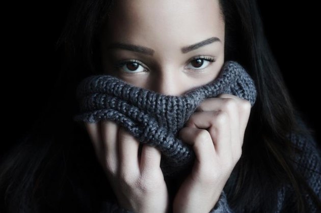 A young woman covers her face with a turtleneck, so that only the eyes and forehead are visible.