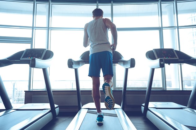 Full-length rear view of young man in sportswear running on treadmill in front of window at gym