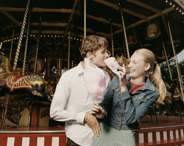 Teenage Couple Stand in Front of a Carousel, Girl Feeding Her Boyfriend Candy Floss and Laughing
