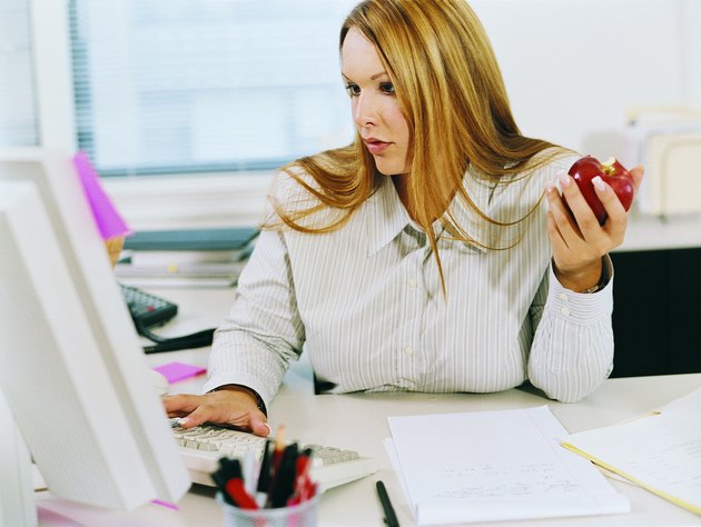 Businesswoman at a Desk Looking at a PC and Eating an Apple