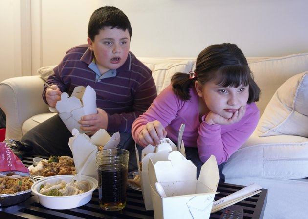 超重的哥哥和姐姐并排坐在沙发上吃着外卖，看着电视