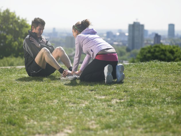 Woman helping man doing sit-ups on grass, city in background