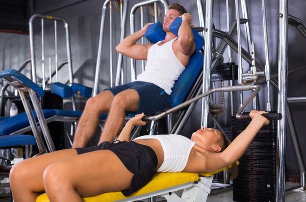 young sporty woman lying on back on bench and lifting weights in shoulder press machinery in gym