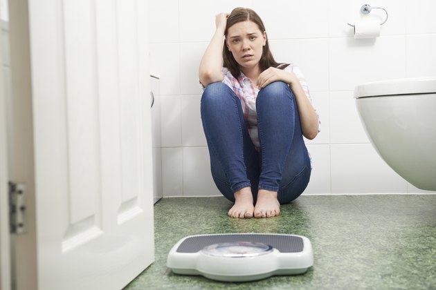 Unhappy Teenage Girl Sitting On Floor Looking At Bathroom Scales