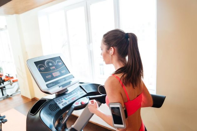 Beautiful serious young woman athlete listening to music from blank screen smartphone and running on treadmill