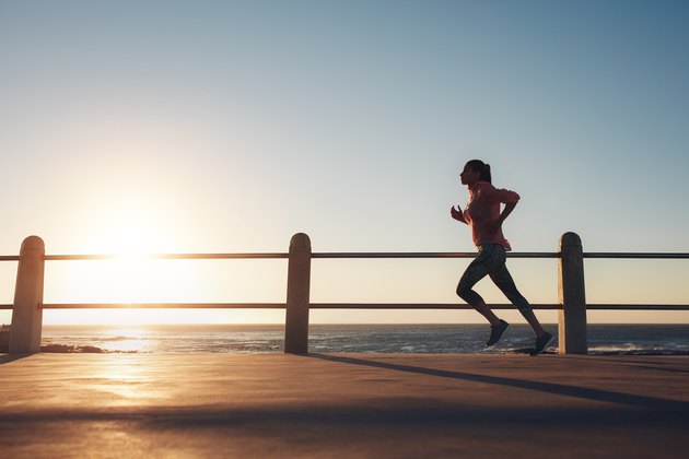 Image of fit sportswoman jogging on a road by the sea during evening. Female runner training at sunset.