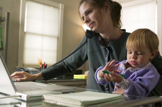a caucasian mother sits working on her computer and talking on the phone with her young daughter on her lap