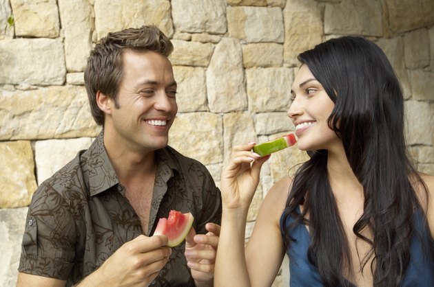Couple eating melon slices outdoors, looking at each other, laughing