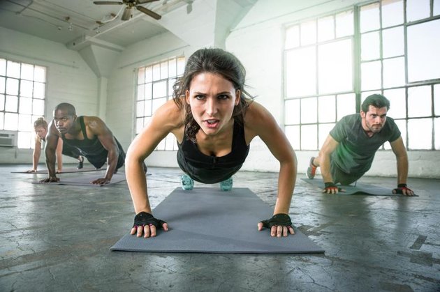 Female leads cross fit power yoga group class strength determination drive as physical trainer instructor in push ups intense focused expression. Fitness exercise gym group muscle building workout with woman leading group of people