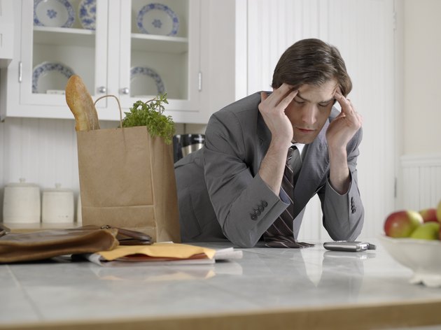 Businessman with bag of groceries rubbing forehead in kitchen