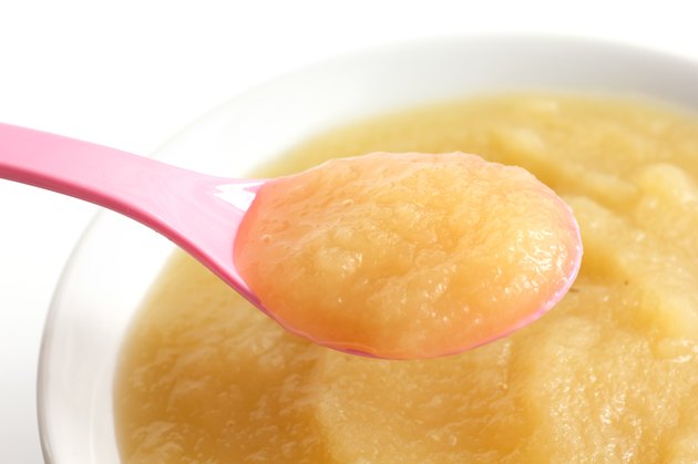 White dish of apple sauce with pink baby spoon.