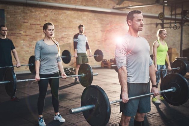 Serious muscular bearded man in shorts and gray tee shirt leading small group of young adults in barbell exercises for fitness training