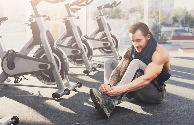 Little break. Handsome young man in sportswear relax after training in gym, sit on floor, lacing sneakers shoes. Sexy guy with tattoo in fitness center