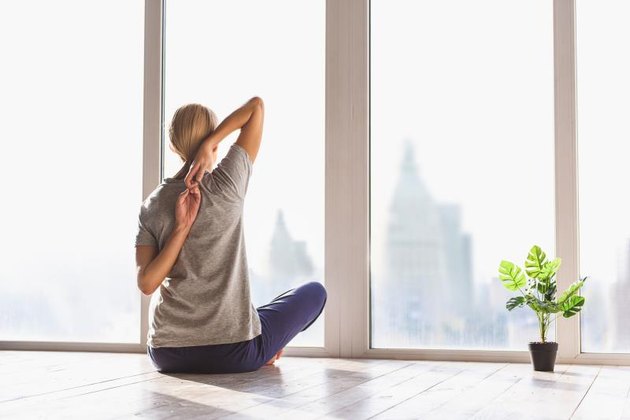 Young woman is undergoing morning exercise. She is sitting and stretching arms behind back. Lady is looking at window and enjoying urban view