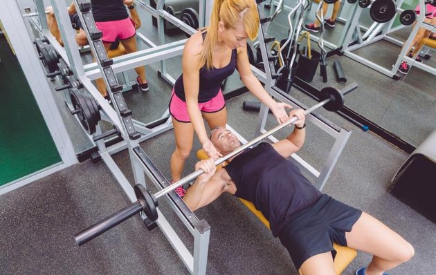 Woman personal trainer helping to muscle man for a correct bench press training with barbell on fitness center