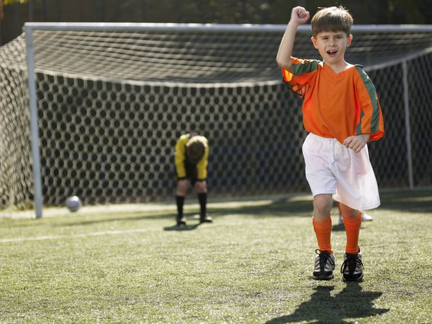 Boy in soccer uniform celebrating, defeated goalkeeper in background (portrait)