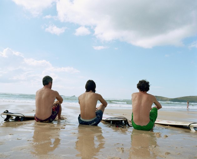 Three men sitting on wet sand by surfboards, rear view