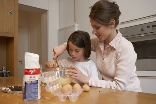 Mother helping daughter (6-8) stir cake mixture in kitchen, smiling