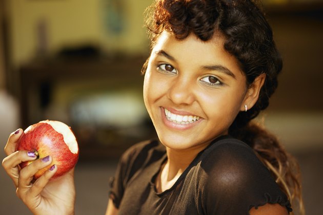 Close-up of a teen girl smiling at the viewer while holding the apple that she is eating.