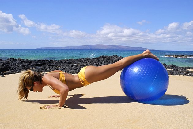 Woman doing pushups with her legs on and exercise ball on a tropical beach.