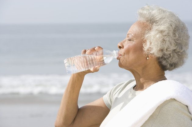 Senior woman stands with drinks water with a towel over her shoulder on beach