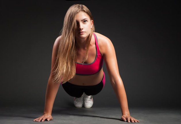 Young girl in sport outfit making push-up on hands and looking at camera on plain black background.
