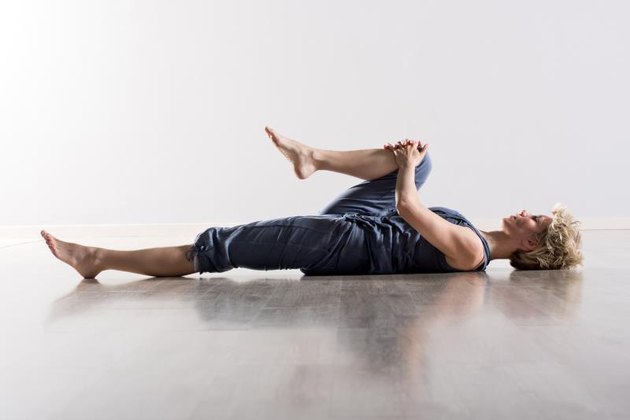 Single athletic woman with blond hair in blue outfit on back stretching hamstring muscles while holding knee close to her chest