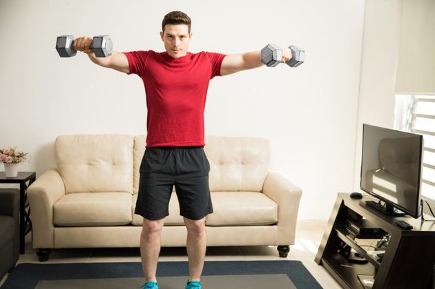 Athletic Hispanic young man exercising his shoulders and doing side raises with a pair of dumbbells at home