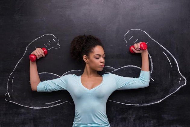 Funny picture of young afro-american woman with dumbbells on chalkboard background. Two strong muscular arms painted on chalkboard