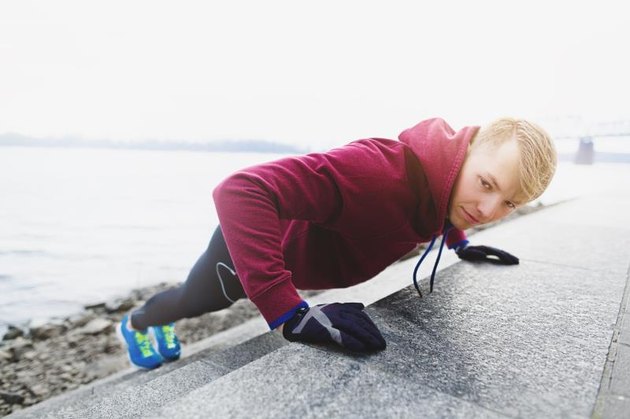 A profile DSLR photo of young male athlete wearing blue running shoes, black tights (pantyhose) and crimson t-shirt with hood. He is doing incline push ups leaning over the stairs of embankment. He is alone in this cold morning. He is looking towards the camera.