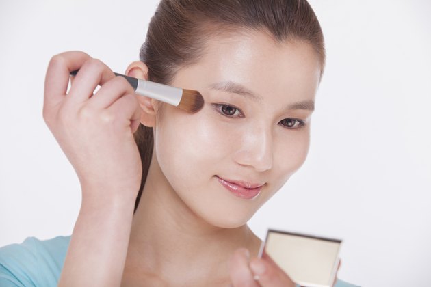 Young beautiful woman looking into a mirror and applying make up with a make up brush, studio shot