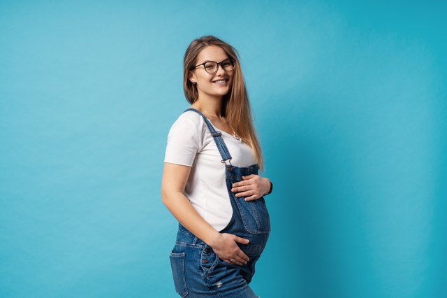 Smiling pregnant woman wearing glasses caressing her belly over blue background