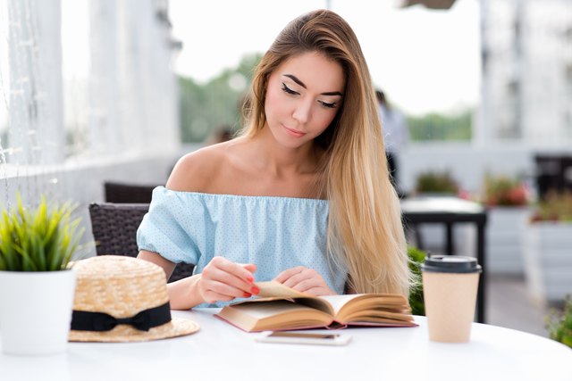 Young and beautiful girl with long hair. On the table is a cup of coffee and tea. Woman reading a paper book on the table. An interesting novel, turns the page. In the summer in the café sun terrace.