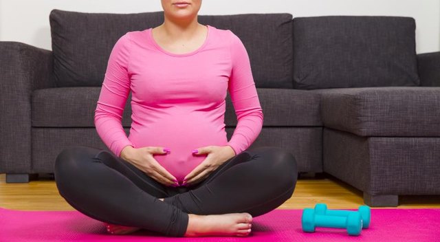 Pregnant woman taking a break from exercising with dumbbells. Sitting on a fitness mat and holding her hands on the swollen belly.