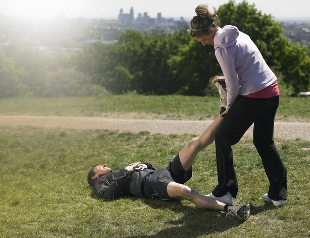 Mid adult man lying on grass after exercising, woman standing doing massage, city in background