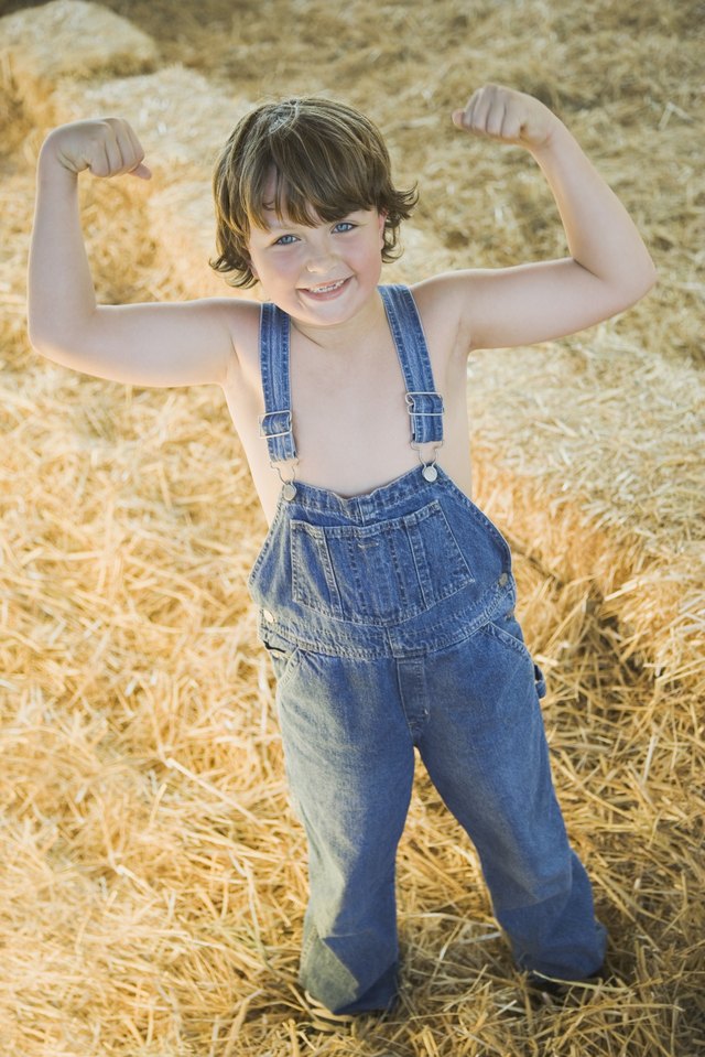 Young boy wearing overalls and flexing muscles in hay