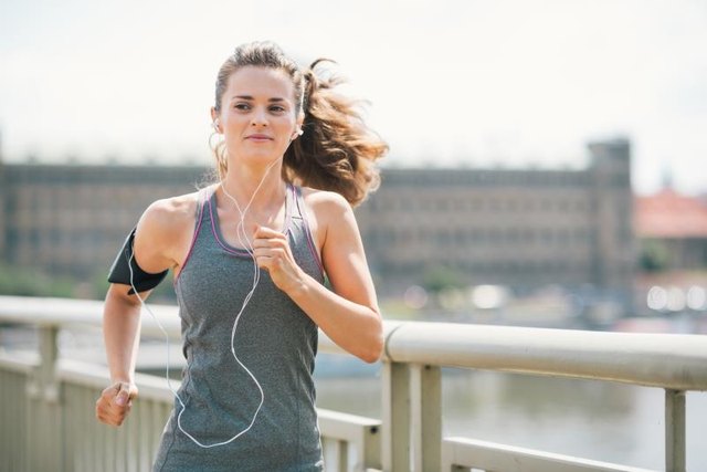 An athletic woman is jogging on a bridge, listening to music. Her long hair is up in a ponytail and blowing in the wind.