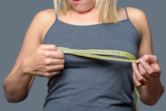 Young blond woman measuring her breast against grey wall Background