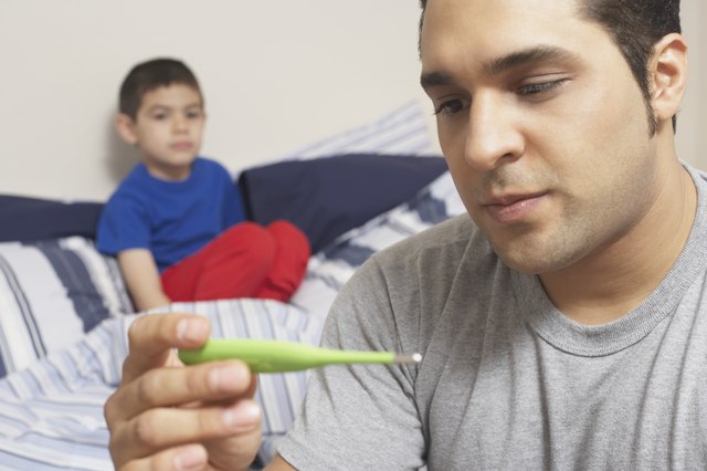 Close-up of a young man holding a digital thermometer with his son sitting on the bed behind him