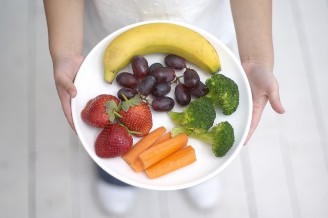 Girl holding plate of fruit and vegetables, including banana, strawberries, grapes, broccoli, carrots