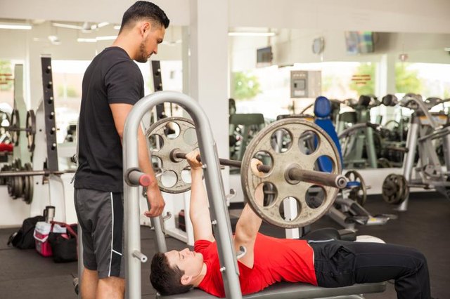 Profile view of a personal trainer spotting a young man while he lifts a barbell on the bench press at a gym