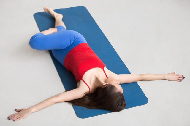 Beautiful happy young woman in bright colorful sportswear working out indoors on blue mat. Girl lying in Reclining Spinal Twist, Jathara Parivartanasana, resting after practice, full length, top view