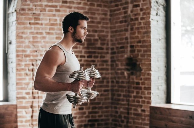 Side view of confident muscled young man wearing sport wear and exercising with dumbbells in loft interior