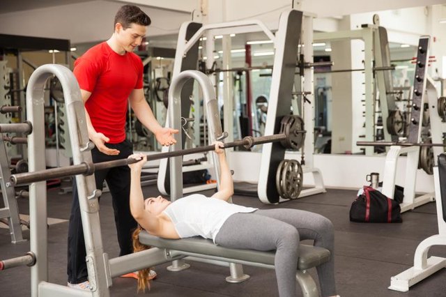 Young athletic woman lifting a barbell on a bench press while her personal trainer spots her at a gym