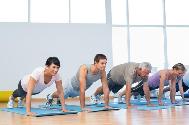 A group of young and old men and women perform push-ups on yoga mats at a sunny gym.