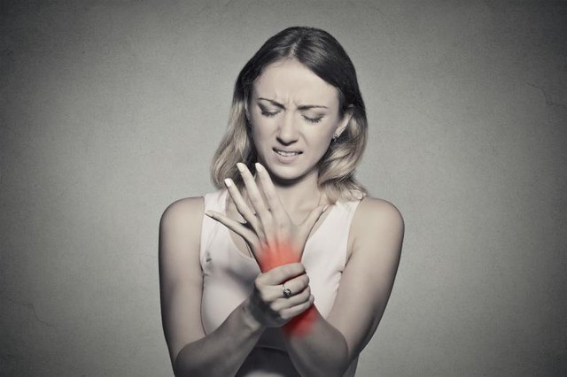 Young woman holding her painful wrist isolated on gray wall background. Sprain pain location indicated by red spot. Negative face expression .