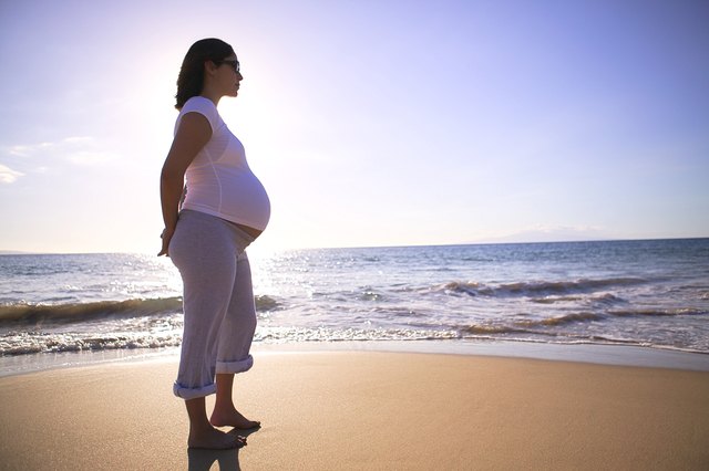 Backlit profile of a pregnant woman standing on the beach near the ocean.
