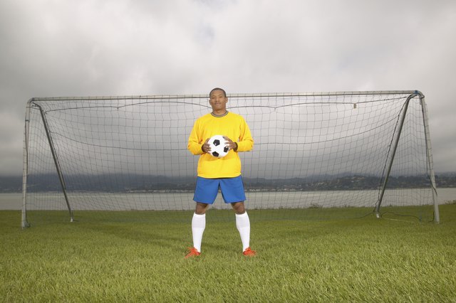Young male football player standing at a goal post holding a soccer ball