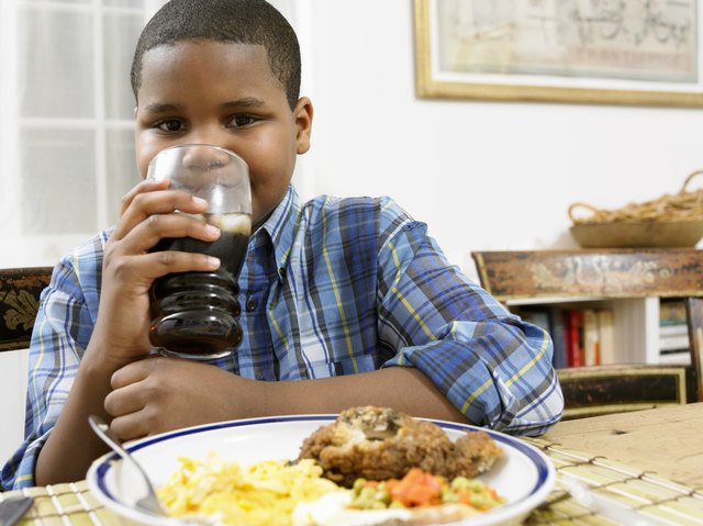 Young Boy Sits at a Table With a Plate of Food, Drinking a Glass of Cola