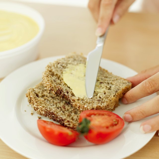 Close-up of a woman's hand putting butter on a slice of brown bread and tomato beside it